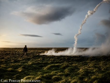 A person stands on grassy terrain near a plume of smoke against a sunset sky.