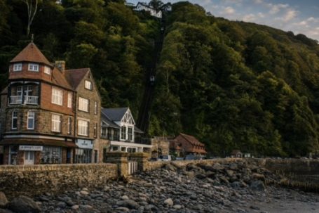 Up And Away - Lynmouth Coastal scene with historic buildings beside a rocky beach and lush green hills.