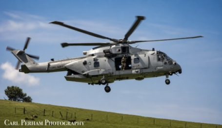 Military helicopter flying over a grassy landscape under a blue sky.