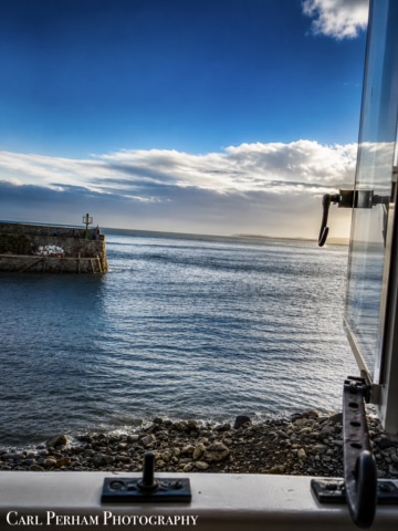 Windowsill And Waves - Clovelly Coastal view featuring a lighthouse, calm sea, and a blue sky with scattered clouds.