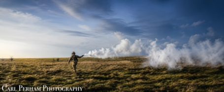 A person walking through a field with smoke near the horizon under a blue sky.