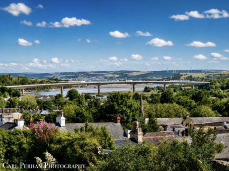 View of a river and bridge surrounded by greenery and blue sky with clouds.