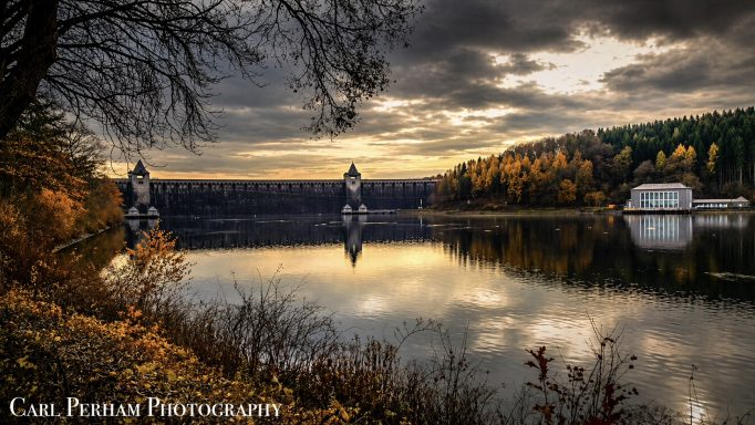 Landscapes From Around The World Serene lakeside view at sunset, with autumn foliage and two water towers reflected in the water.