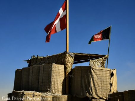 Danish and Afghan flags flying above a sandbagged military outpost in a desert setting.