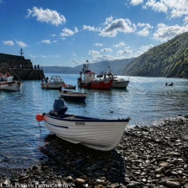 Clovelly A small boat on a rocky shore with fishing boats in the background and hills nearby.