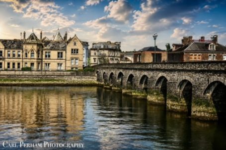 Historic buildings along a riverbank, with arches reflected in the water under a cloudy sky.