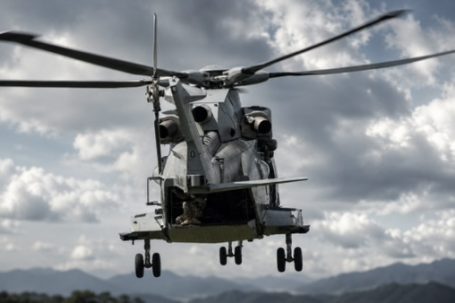 Military helicopter in flight against a cloudy sky, with mountains in the background.