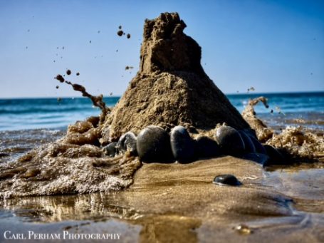 Sea Always Wins - Westward Ho! A sandcastle on a beach with waves splashing around it under a clear blue sky.